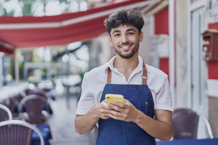 Mesero sonriendo mientras escribe en su teléfono en la terraza de un restaurante